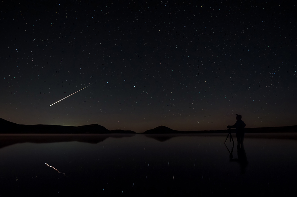 Cena noturna de fotógrafo observando meteoro passando no céu.