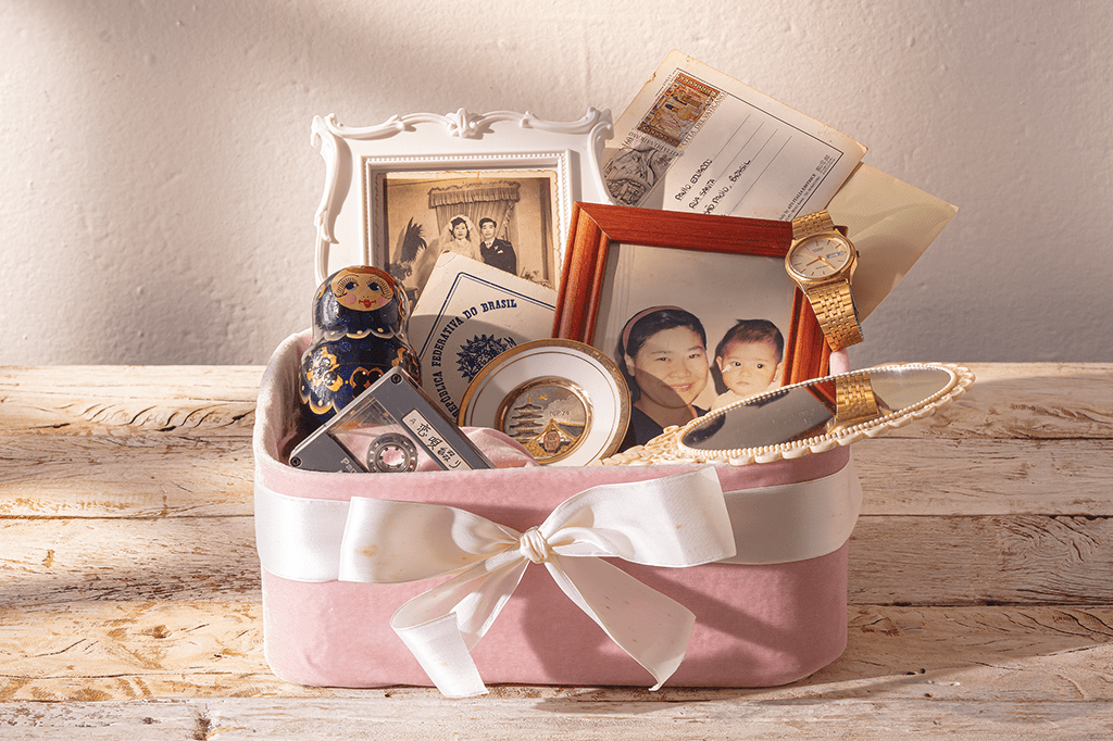 SI_435_Alzheimer_01 Foto de uma caixa de veludo rosa, com um laço em volta. Têm vários objetos dentro dela: um espelho, dois retratos, cartas, uma certidão de nascimento, um pratinho decorativo, uma matriosca, um relógio e uma fita cassete.