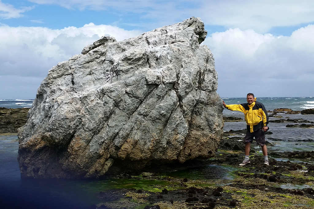 SI_Terremoto_1 Imagem de um homem, em pé, apoiando a mão direita em uma grande rocha, perto do mar.