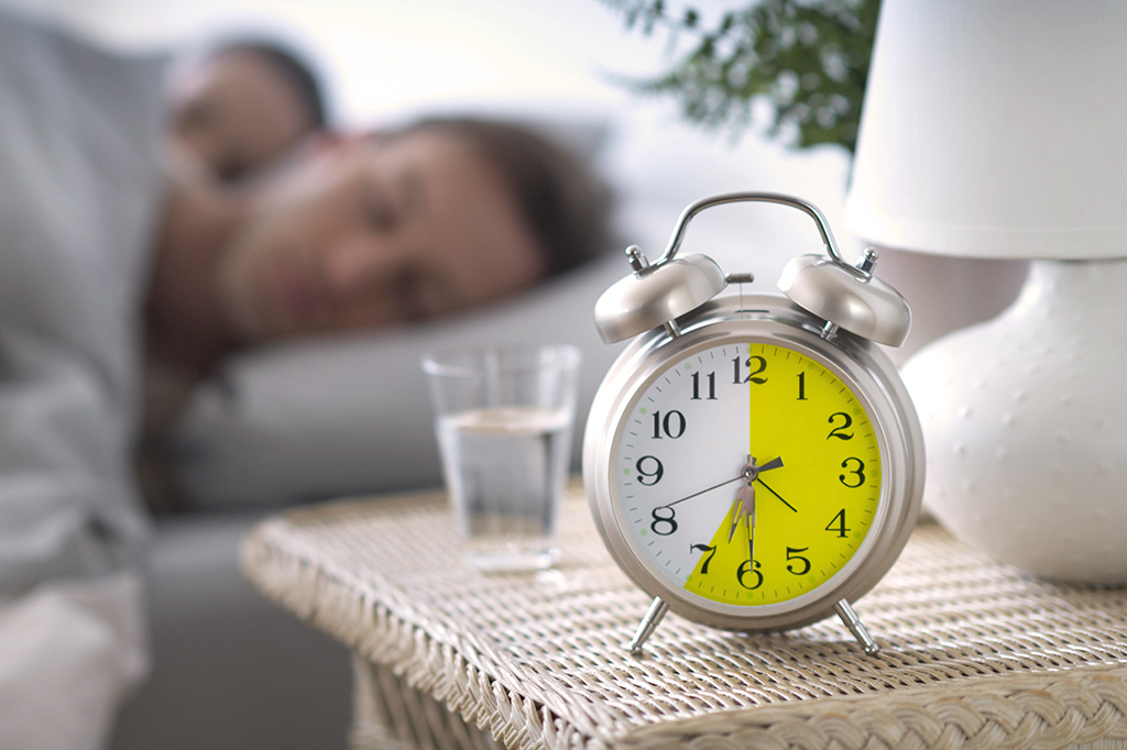 Foto de um casal desfocado e dormindo e, na frente, um relógio com destaque para a marcação de horário entre 12h e 7h.