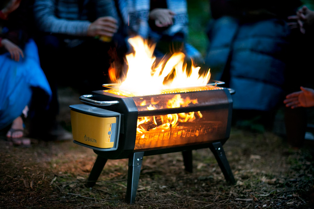 Churrasqueira BioLite FirePit+ acesa, ao ar livre, com pessoas em volta.