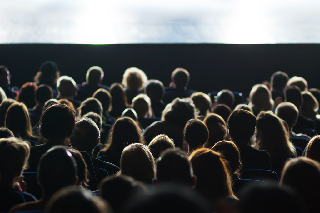 Silhueta de pessoas de costas, sentadas, assistindo filme em sala de cinema.