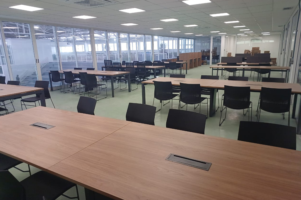 Sala de aula com diversas mesas que possuem 8 cadeiras cada. A sala está no segundo andar do edifício. As paredes são de vidro.