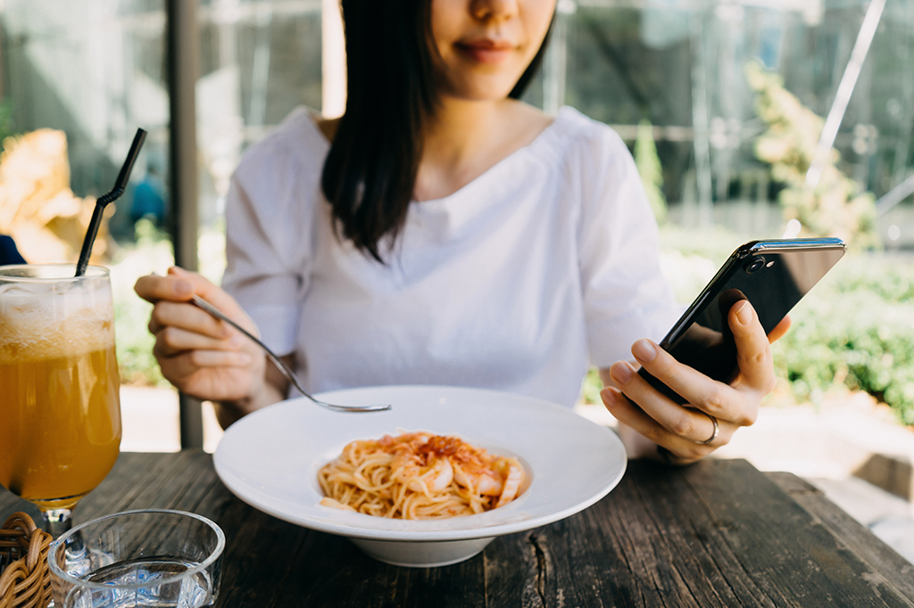 Uma mulher segurando o celular em sua mão esquerda e um garfo em sua mão direita. Ela está sentada em uma mesa, a qual possui um prato de comida e uma bebida ao lado.