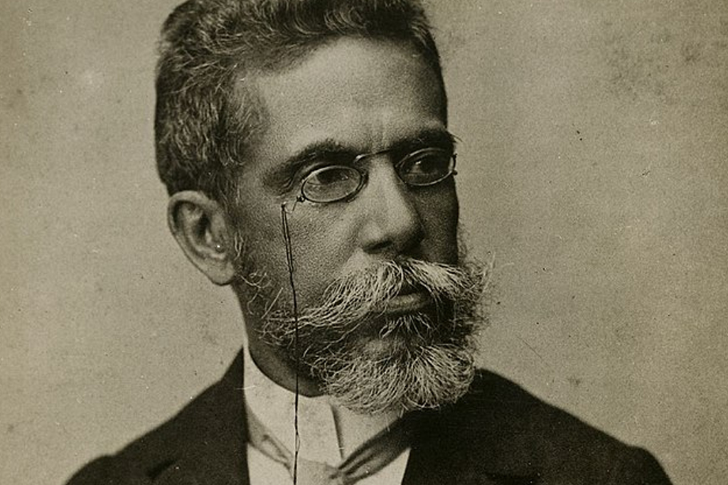Machado de Assis. Na foto, o homem veste um par de óculos. Possui uma barba com fios brancos.
