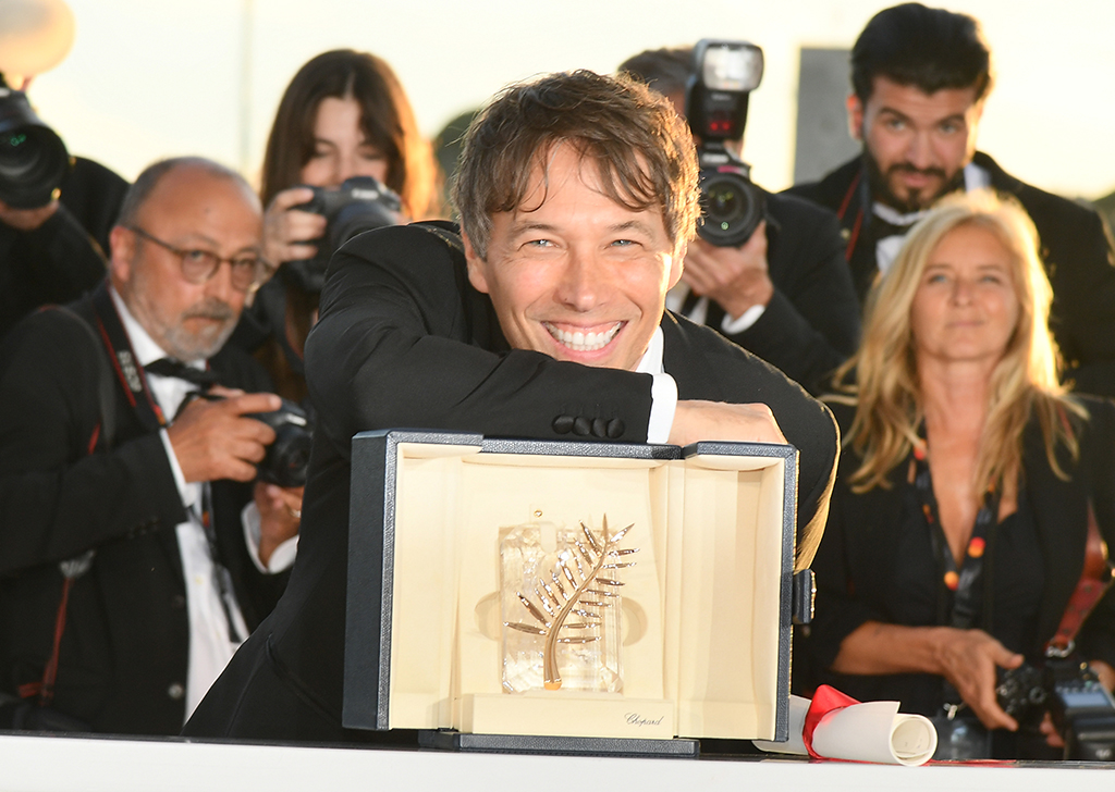 Um homem vestido de terno com a mão e o rosto apoiados em cima de uma caixa, que possui um prêmio em seu interior. Há fotógrafos ao fundo.