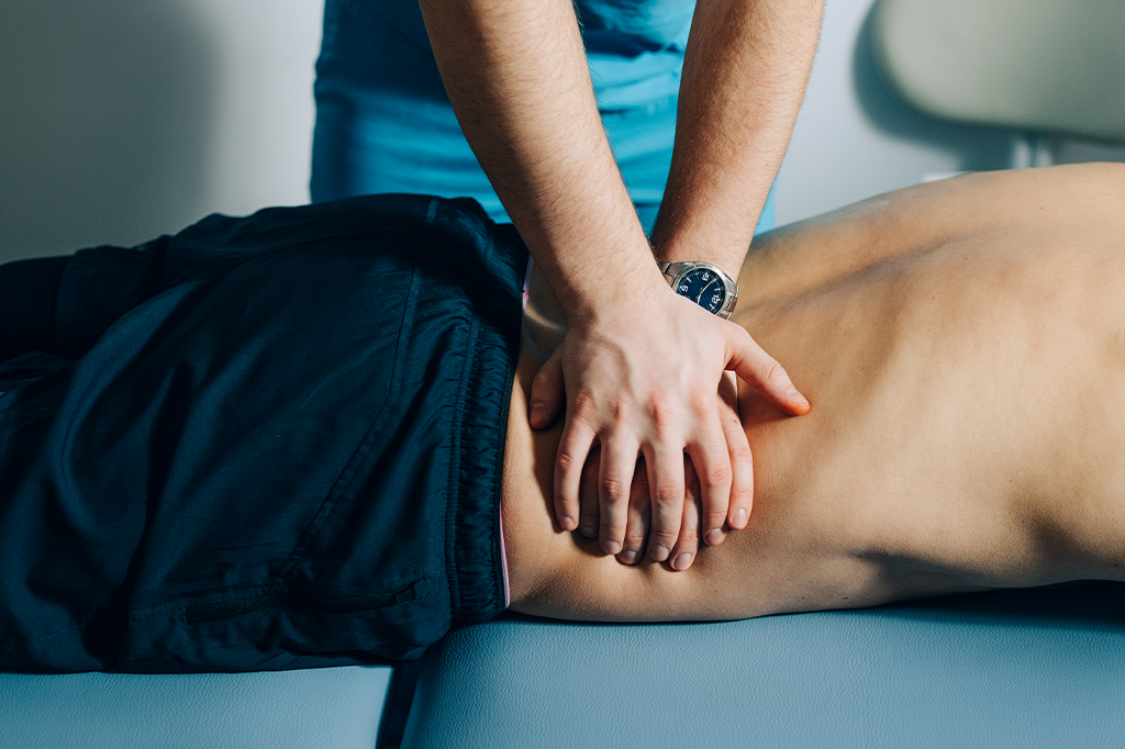 Uma pessoa debruçada em uma maca, com um quiropraxista fazendo massagem em suas costas.