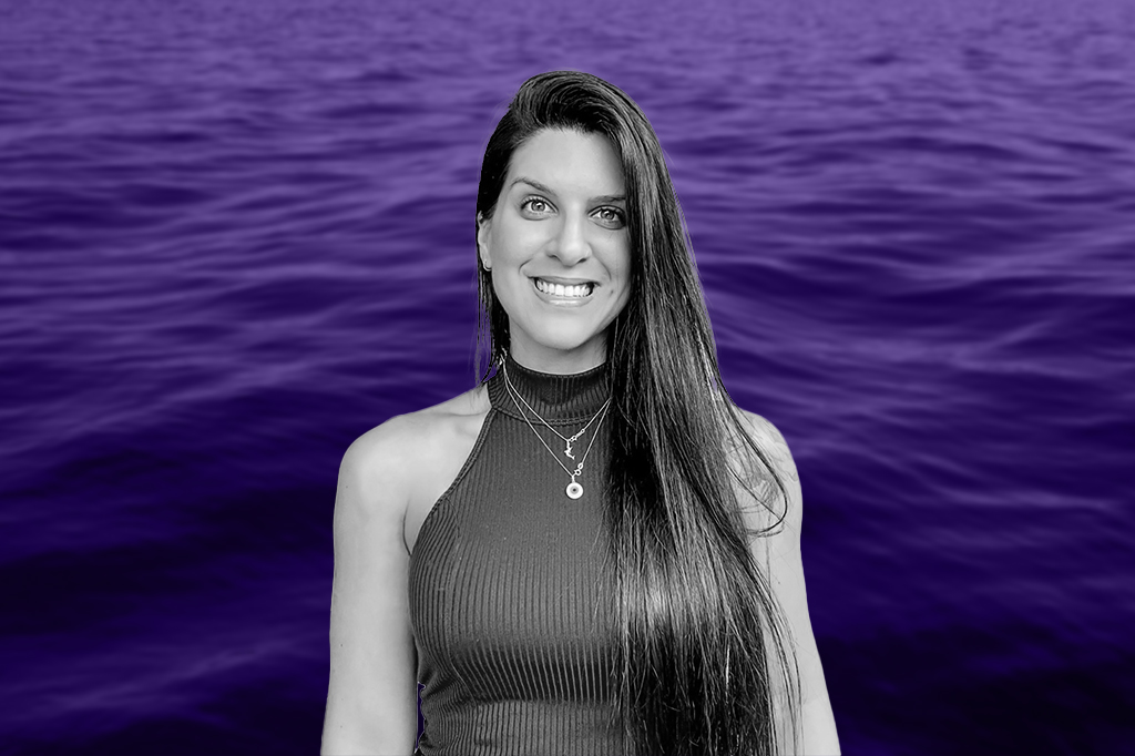 Kamilla Souza. Mulher da cabelos cumpridos e blusa gola alta sem manga. Está sorrindo para a foto. No fundo, há um oceano.