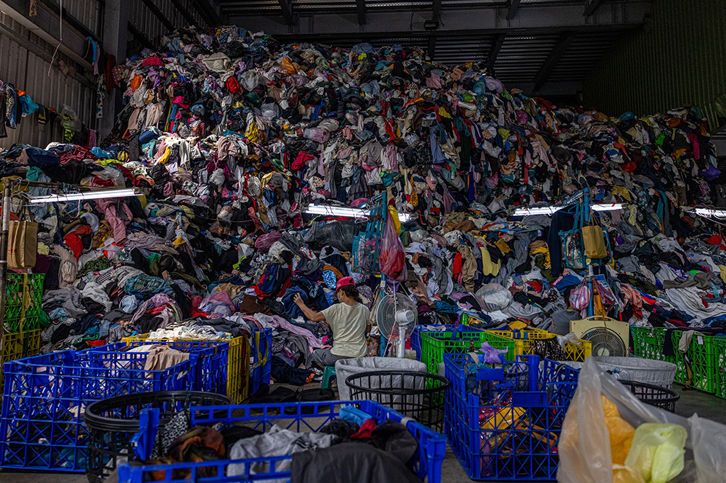 Fábrica de reciclagem têxtil com enorme pilhagem de tecidos. Há uma mulher vasculhando-os.