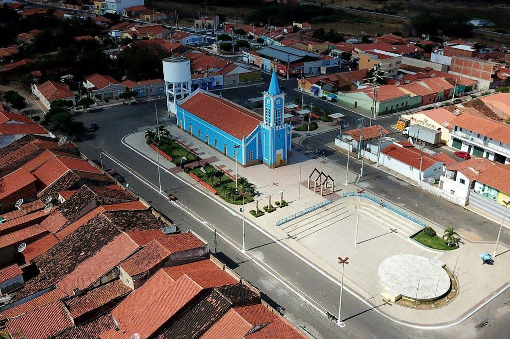 Visão aérea de uma cidade. Há diversas casas e uma igreja e uma praça, ao centro.