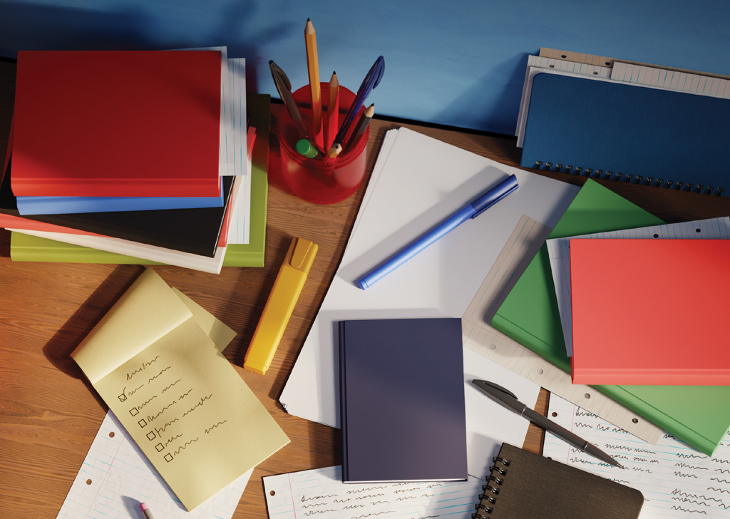 Imagem de uma mesa bagun&ccedil;ada, com muitos cadernos, pap&eacute;is e canetas espalhados.