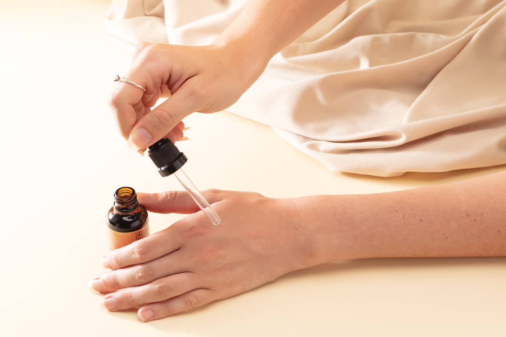 Imagem focada nas m&atilde;os de uma mulher aplicando um rem&eacute;dio em gotas na pele.