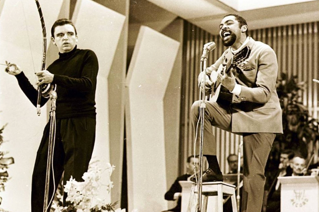 Imagem de um rapaz, vestido de preto, tocando um berimbau. A direita, um rapaz de terno marrom, cantando e tocando viol&atilde;o. Cena do filme uma noite em 67 (2010).