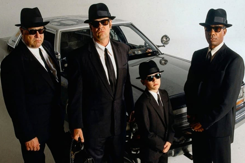 Imagem de tr&ecirc;s adulto e uma crian&ccedil;a vestidos de terno preto, chap&eacute;u e &oacute;culos escuros. Ao fundo, um grande autom&oacute;vel. Cena do filme Blues Brothers 2000 (1998).